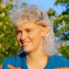 Eine junge Frau mit lockigem, weissblondem Vokuhila-Haarschnitt, die an einem sonnigen Tag draussen sitzt.