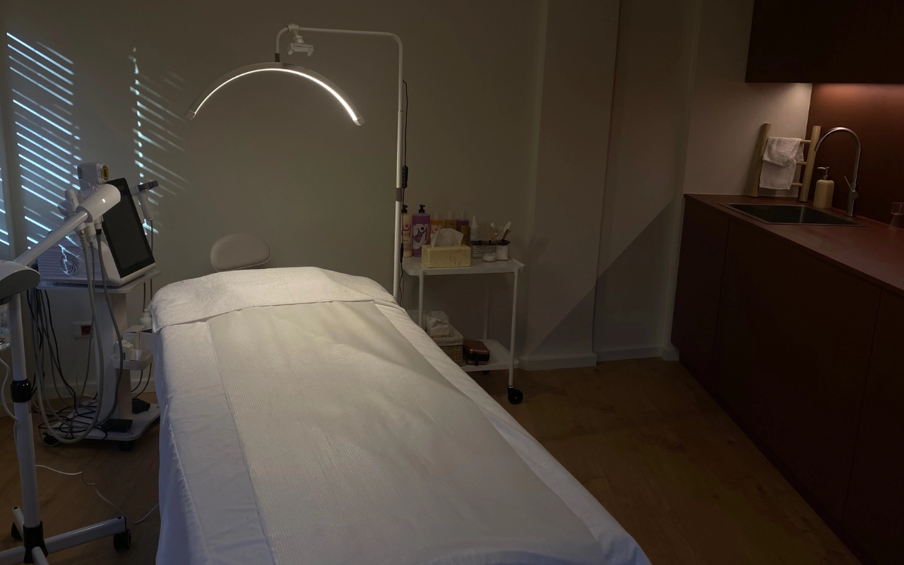 Ein konzentrierter Behandlungsraum mit weissem Bett unter einer Bogen-LED-Lampe, professioneller Ästhetik-Ausrüstung mit digitalem Bildschirm, Hautpflegeprodukten auf einem Trolley und dunklen Holzschränken mit Waschbecken an der hinteren Wand.
