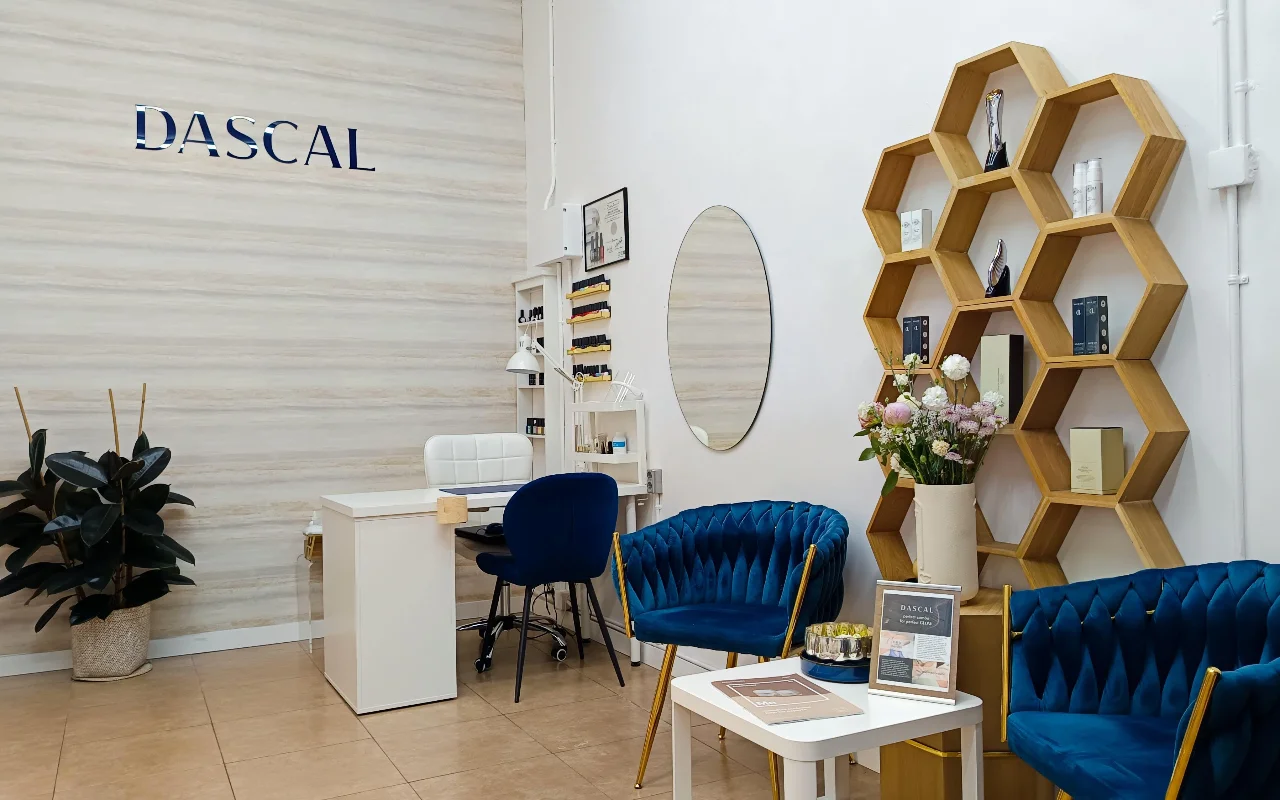 Ein stilvolles Beauty-Salon-Interieur mit tiefblauen Samtsesseln und einem sechseckigen Holzregal an der Wand. Ein weisser Maniküretisch steht in der Ecke an einer hellen, texturierten Wand mit dem Schriftzug DASCAL in blauen Buchstaben.