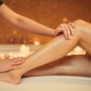 Seitenansicht der Hände einer Kosmetikerin, die sanft über die Beine einer Frau streichen, während diese nach einem Waxing auf einer Behandlungsliege liegt