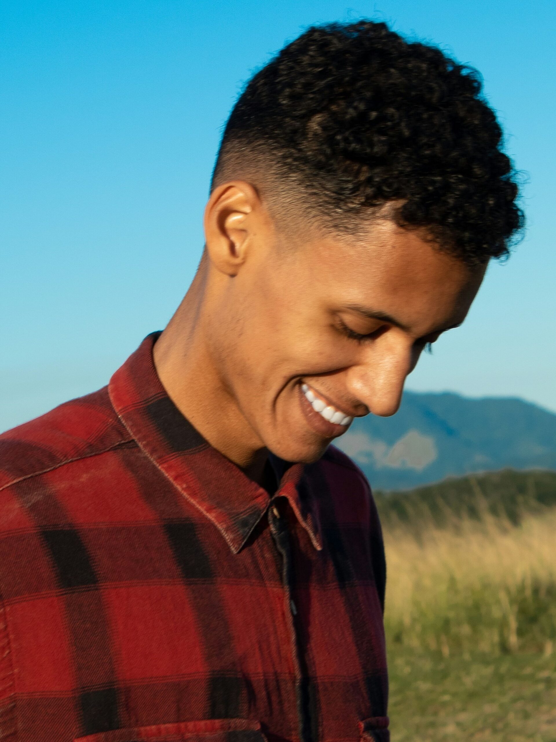 Lächelnder Mann im Freien vor Bergen und blauem Himmel mit lockigem Haar und einem Mid Taper Fade