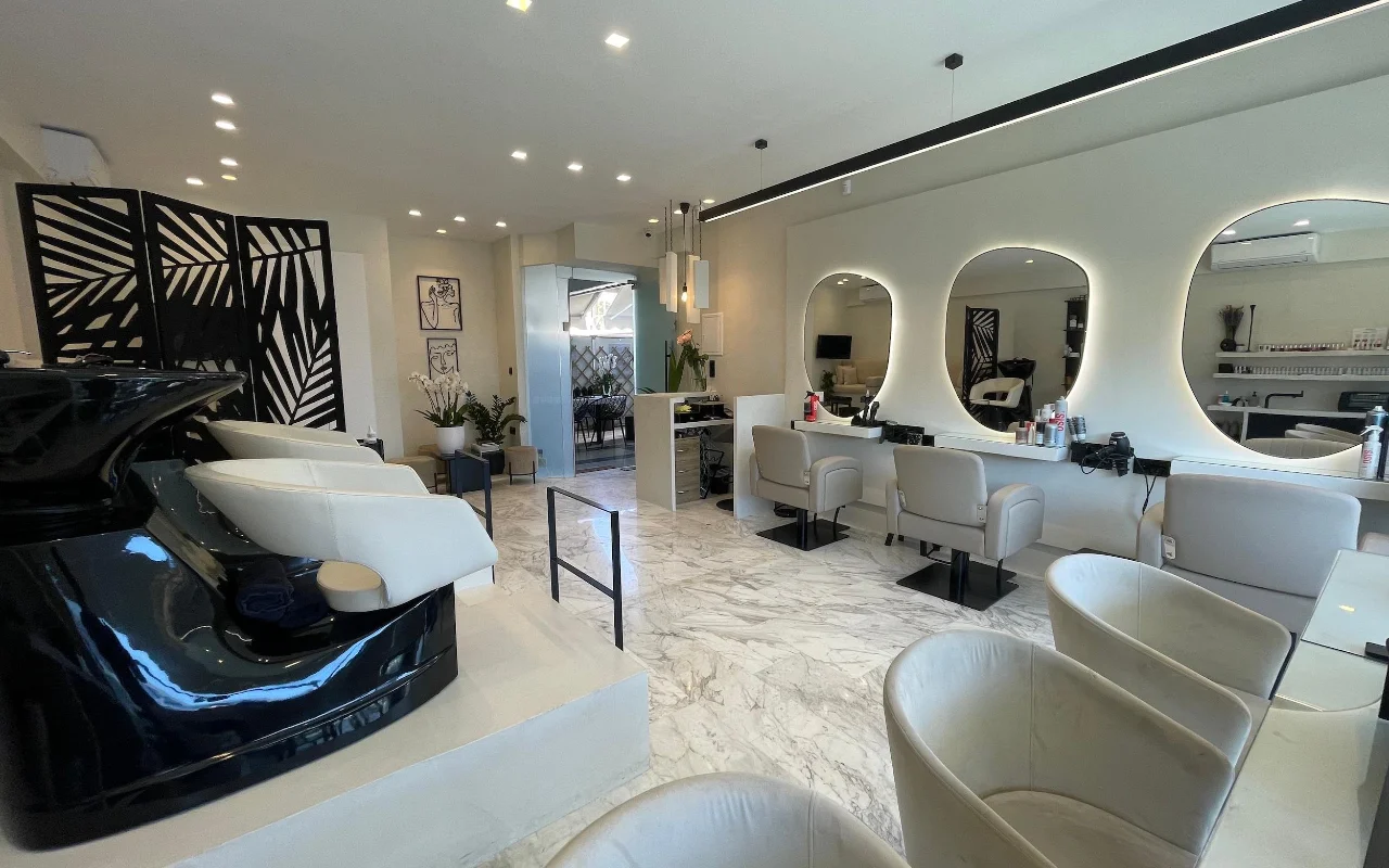 Ein eleganter Friseursalon mit weissem Marmorboden, cremefarbenen Leder-Stylingstühlen unter LED-gerahmten Ovalspiegeln, einer glänzenden schwarzen Waschstation und einem geometrischen Palmblatt-Raumteiler.