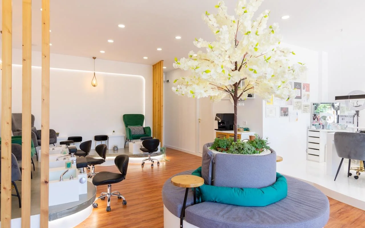 Ein offenes, naturinspiriertes Nagelstudio mit einem grossen dekorativen weissen Blütenbaum als Mittelpunkt, smaragdgrünen Samtfauteuils an den Pedikürestationen, Holzlamellentrennwänden und warmem Holzboden.