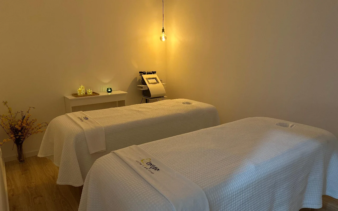 Ein ruhiger Massage- oder Behandlungsraum mit zwei Behandlungsliegen nebeneinander, bezogen mit weissen Handtüchern und Laken. Der Raum wird von einer einzelnen warmen Hängeglühbirne und kleinen Kerzen auf einem Beistelltisch beleuchtet und schafft eine entspannte Atmosphäre. Ein spezialisiertes Kosmetikgerät steht im Hintergrund zwischen den Liegen.