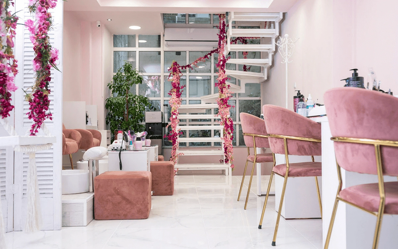 Ein feminines Nagelstudio mit zartrosa Samtsesseln, einer weissen Wendeltreppe mit rosa Kunstblumen, Lamellentrennwänden und goldbeinigen Manikürestationen auf marmorähnlichem Boden.