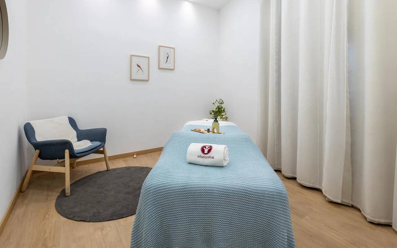 Ein sauberer, minimalistischer Spa-Raum mit weissen Wänden und hellem Holzboden. Eine Behandlungsliege ist ordentlich mit einer hellblauen Strickdecke und einem gerollten weissen Handtuch mit dem Logo „Vitaforma“ bezogen. In der Ecke steht ein stilvoller blauer Sessel auf einem kleinen runden grauen Teppich, und lange weisse Vorhänge hängen am Fenster.