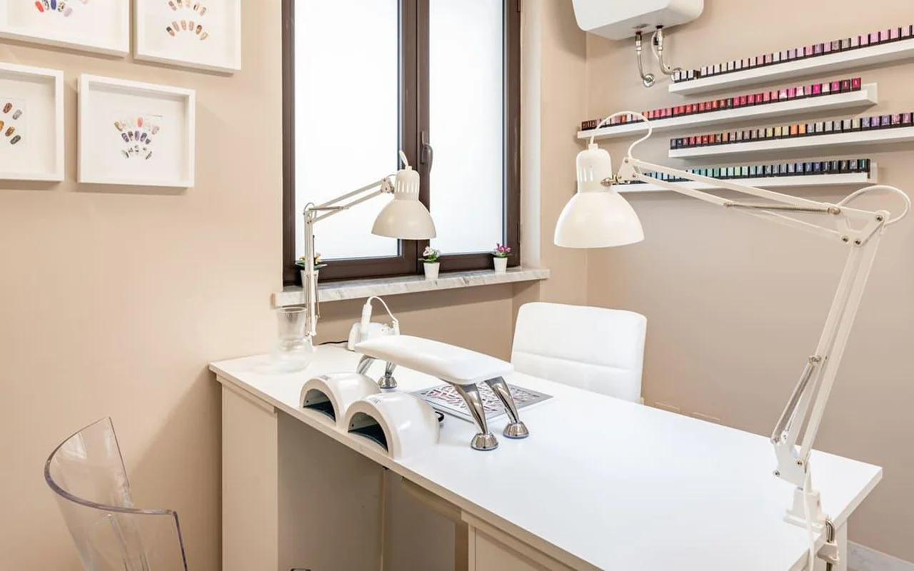 Eine eigene Nagelstation bei AG The Beauty Room's mit einem sauberen weissen Tisch, zwei UV-Lampen, verstellbaren Arbeitsleuchten und Regalen voller bunter Nagellacke. Gerahmte Nailart-Designs hängen an den warmtonigen Wänden.