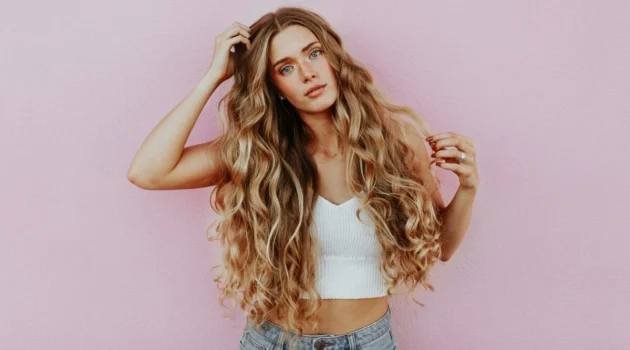 Studioaufnahme einer Frau mit sehr langem, welligem, sonnengeküsstem Balayage-Haar, die eine hellblaue Jeans und ein weisses Strick-Crop-Top trägt
