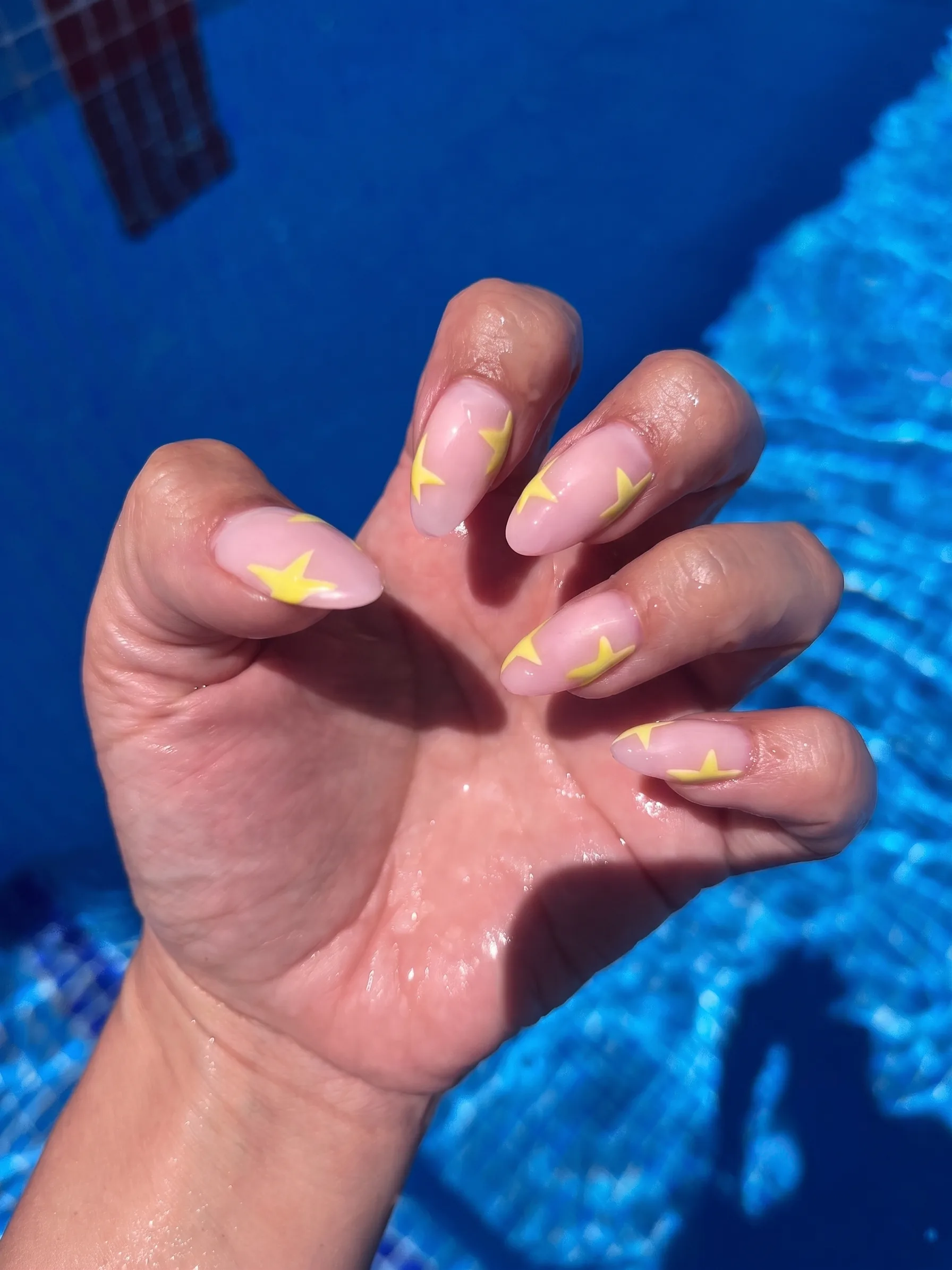 Mandelförmige Nägel in transparentem Nude mit buttergelben Sternen, am Pool fotografiert.