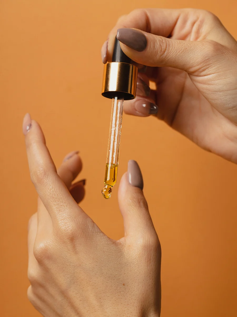 Nahaufnahme zweier gepflegter Hände, eine hält eine Glaspipette, aus der Nagelöl tropft