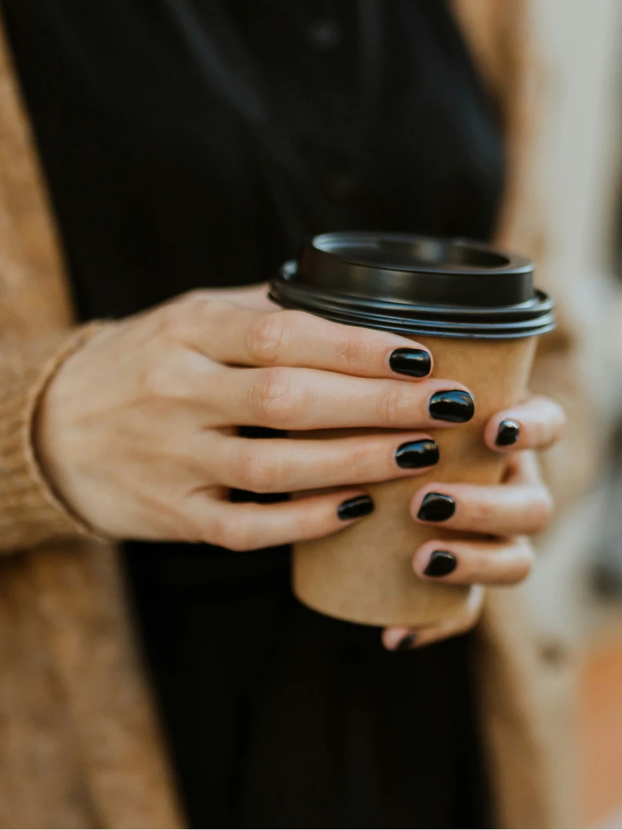 Nahaufnahme einer Frau mit glänzender schwarzer Gel-Maniküre, die einen Coffee-to-go-Becher hält