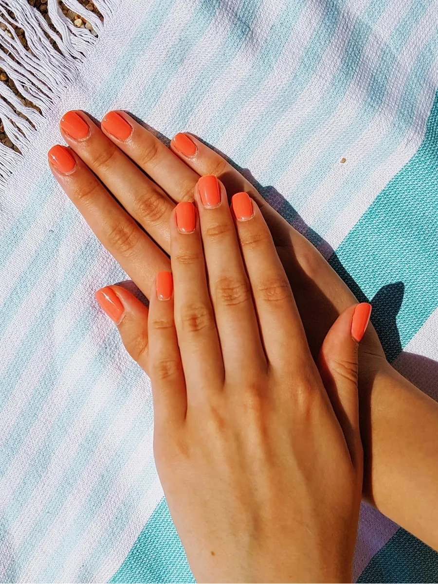 Aufsicht auf zwei Hände mit glänzender oranger Gel-Maniküre auf einem blau-weiss gestreiften Strandtuch