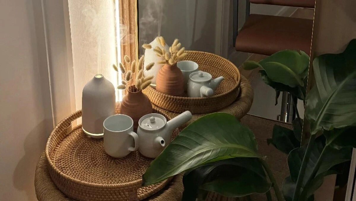 Nahaufnahme eines Korbtabletts mit einem Diffuser und einem Teeservice neben einer grossen grünen Pflanze in einem Salon