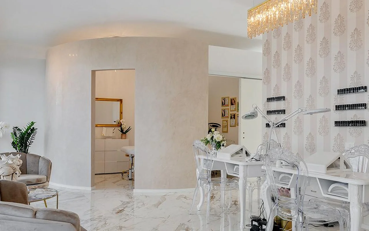 Das elegante Interieur des MC Beauty Center mit Boden in Marmoroptik, weissen geschwungenen Wänden und einem goldenen Kristall-Kronleuchter. Rechts steht eine Nagelstation mit Ghost-Chairs, durch eine goldgerahmte Tür gelangt man in einen Behandlungsraum dahinter.