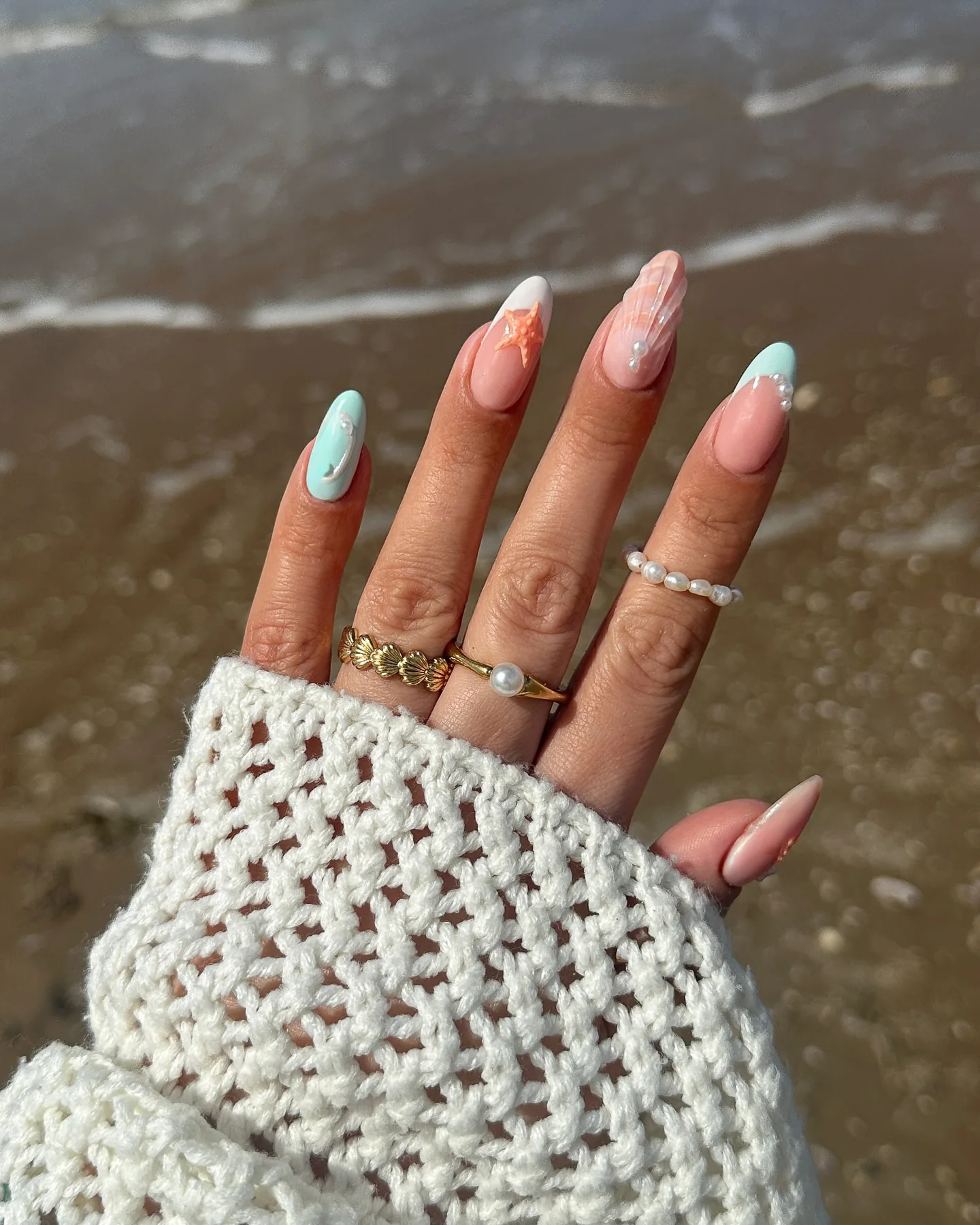 Mandelförmige Nägel mit pastelligem Meerjungfrau-Nail-Art – Seestern, Perlmuttakzente und schillernder Schimmer – auf feuchtem Sand.