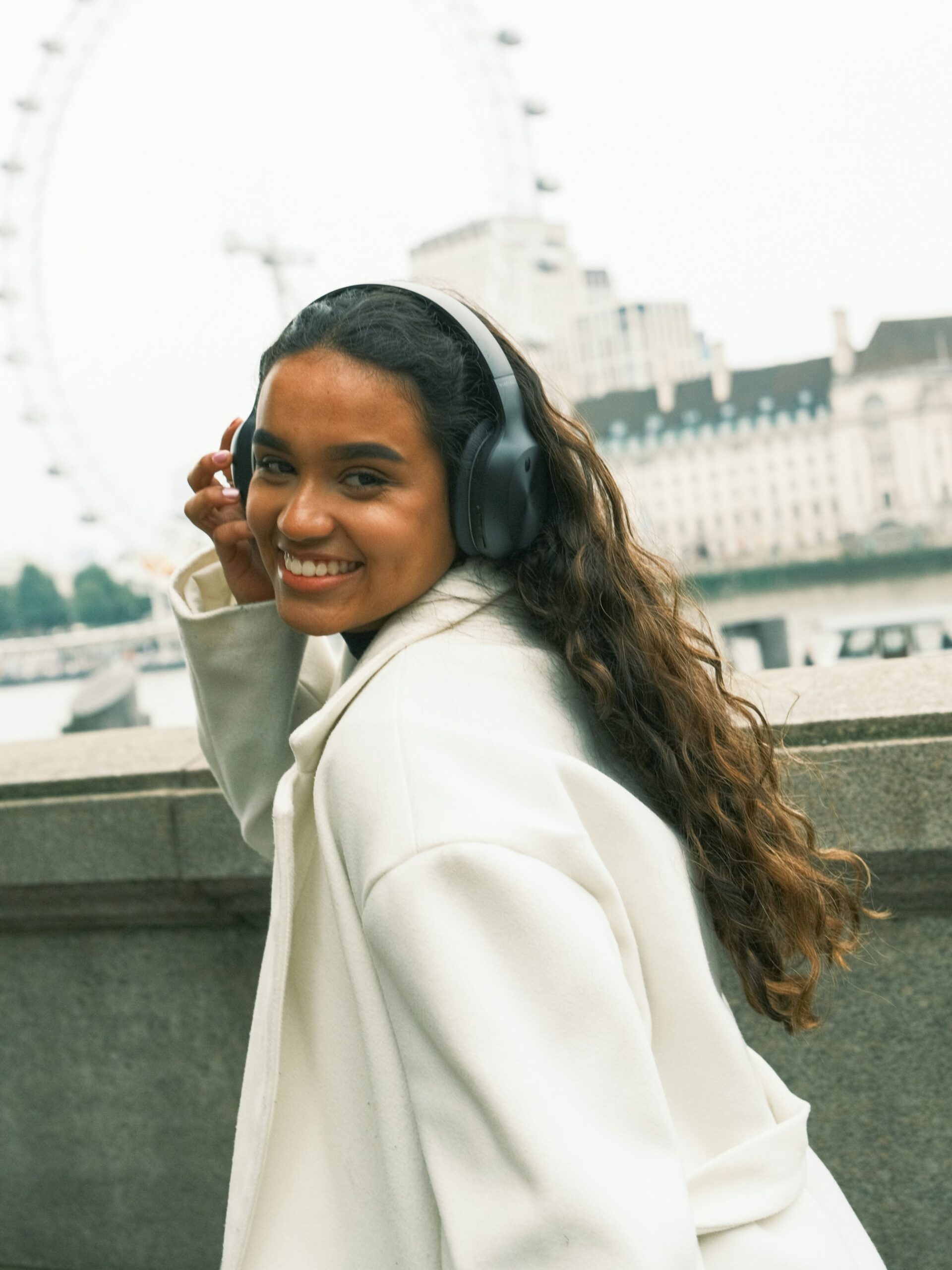 Eine lächelnde Frau mit Microblading an den Brauen, schwarzen Over-Ear-Kopfhörern und einem weissen Mantel auf einer Brücke