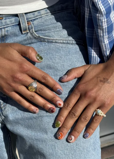 Mismatched Maniküre auf kurzen Nägeln, jeder Nagel in einer anderen Farbe und einem anderen Design, mit rotem Stern und oranger Schnörkellinie, auf Jeans abgelegt.