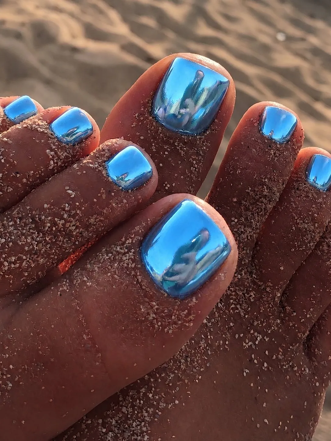 Metallisch glänzende Chrome-Pediküre in Ozeanblau auf sandigen Füssen am Strand