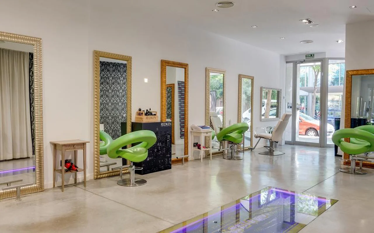 Ein heller, geräumiger Coiffeursalon mit poliertem Betonboden, hohen goldgerahmten Spiegeln an einer weissen Wand, limettengrünen geformten Styling-Stühlen und einem Glasboden-Bereich mit lila Licht, mit Blick auf die Strasse durch verglaste Türen im Hintergrund.