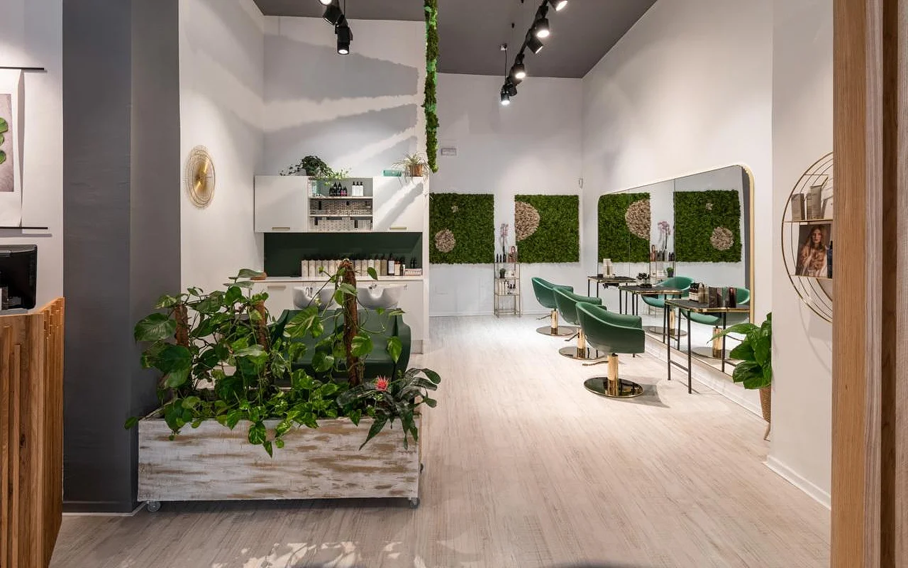 Ein pflanzengefüllter Coiffeursalon mit hellem Holzboden, einem gealterten weissen Übertopf voller Grün, grüner Moos-Wandkunst, smaragdgrünen Samt-Styling-Stühlen mit Golddetails und einer Waschstation dahinter.