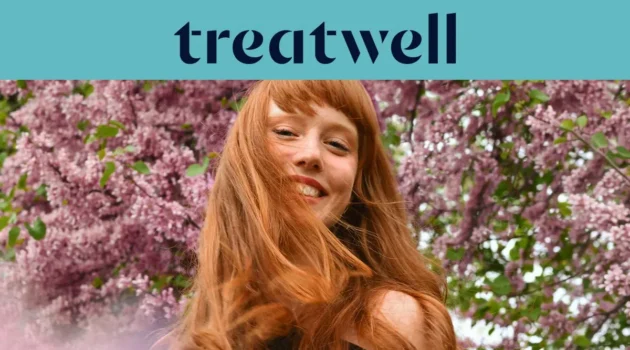 Nahaufnahme einer lächelnden jungen rothaarigen Frau mit Pony, deren Haare vor einem Hintergrund aus rosa Fliederblüten im Wind wehen. Ein hellblaues Banner am oberen Rand zeigt das &bdquo;Treatwell“-Logo in Dunkelblau.