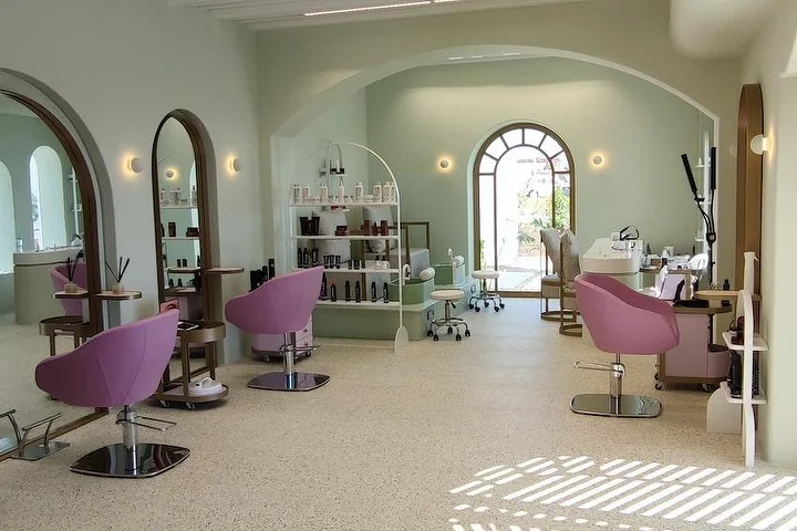 Ein heller, salbeigrüner Salon mit Terrazzoboden, bogenförmigen Spiegeln mit Holzrahmen und einem Bogenfenster zum Garten, altrosa Stylingstühlen und offenen Regalen voller Produkte, während Sonnenlicht über den Boden fällt.