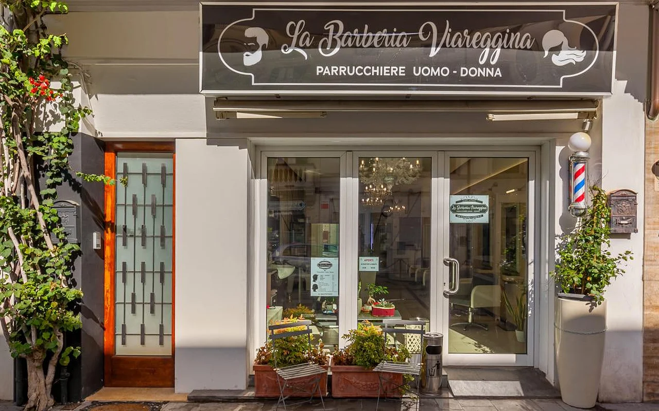 Der Eingang von La Barberia Viareggina an einem sonnigen Tag, mit einer klassischen rot-weiss-blauen Barber-Säule rechts neben den Glastüren. Topfpflanzen und rankendes Grün rahmen den Eingang, und durch die Fenster ist im Inneren ein Kronleuchter zu sehen.