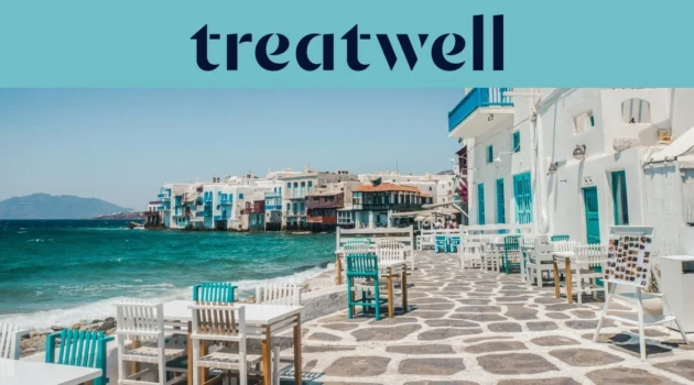 Die Uferpromenade von Klein-Venedig in Mykonos-Stadt: weiss getünchte Häuser mit bunten Holzbalkonen direkt am Wasser, dahinter die türkisfarbene Ägäis und im Vordergrund weisse und petrolfarbene Café-Tische auf einer steingepflasterten Terrasse.