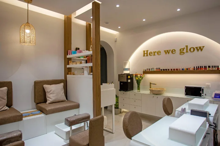 Ein warmer, sanft beleuchteter Nagelsalon mit einer geflochtenen Rattan-Hängelampe, gepolsterten braunen Pediküre-Bänken, weissen Maniküre-Tischen, einem Regal voller bunter Lacke und einem goldenen „Here we glow“-Schriftzug auf einem hinterleuchteten Bogen.