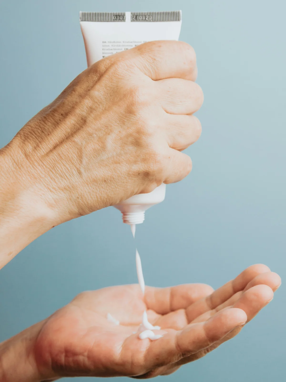 Nahaufnahme einer Hand, die eine Tube mit weisser Feuchtigkeitscreme senkrecht hält und etwas davon in eine darunterliegende Hand drückt