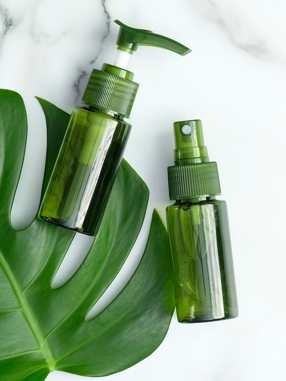 Flatlay mit zwei grünen Fläschchen kühlender Gele und Seren, die auf einem grünen Blatt auf weissem Marmorhintergrund liegen