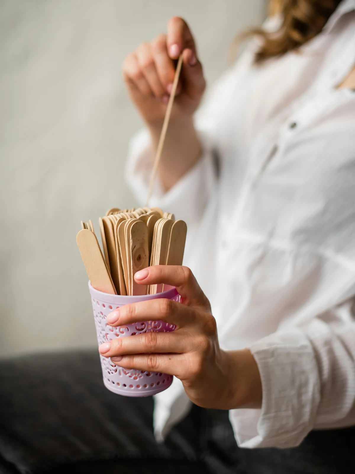 Nahaufnahme einer Frau in einem weissen Shirt, die mit einer Hand ein rosafarbenes Gefäss mit Holzspateln hält und mit der anderen einen Spatel herauszieht