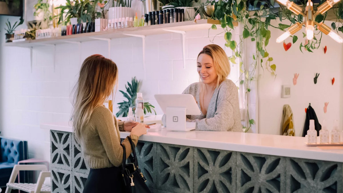 Eine Frau steht an einem Salontresen, während eine Rezeptionistin ihren nächsten Termin bucht; der Salon ist aufgeräumt, mit weissem Tresen, ordentlichen Produktregalen sowie Pflanzen und Blumen als Deko