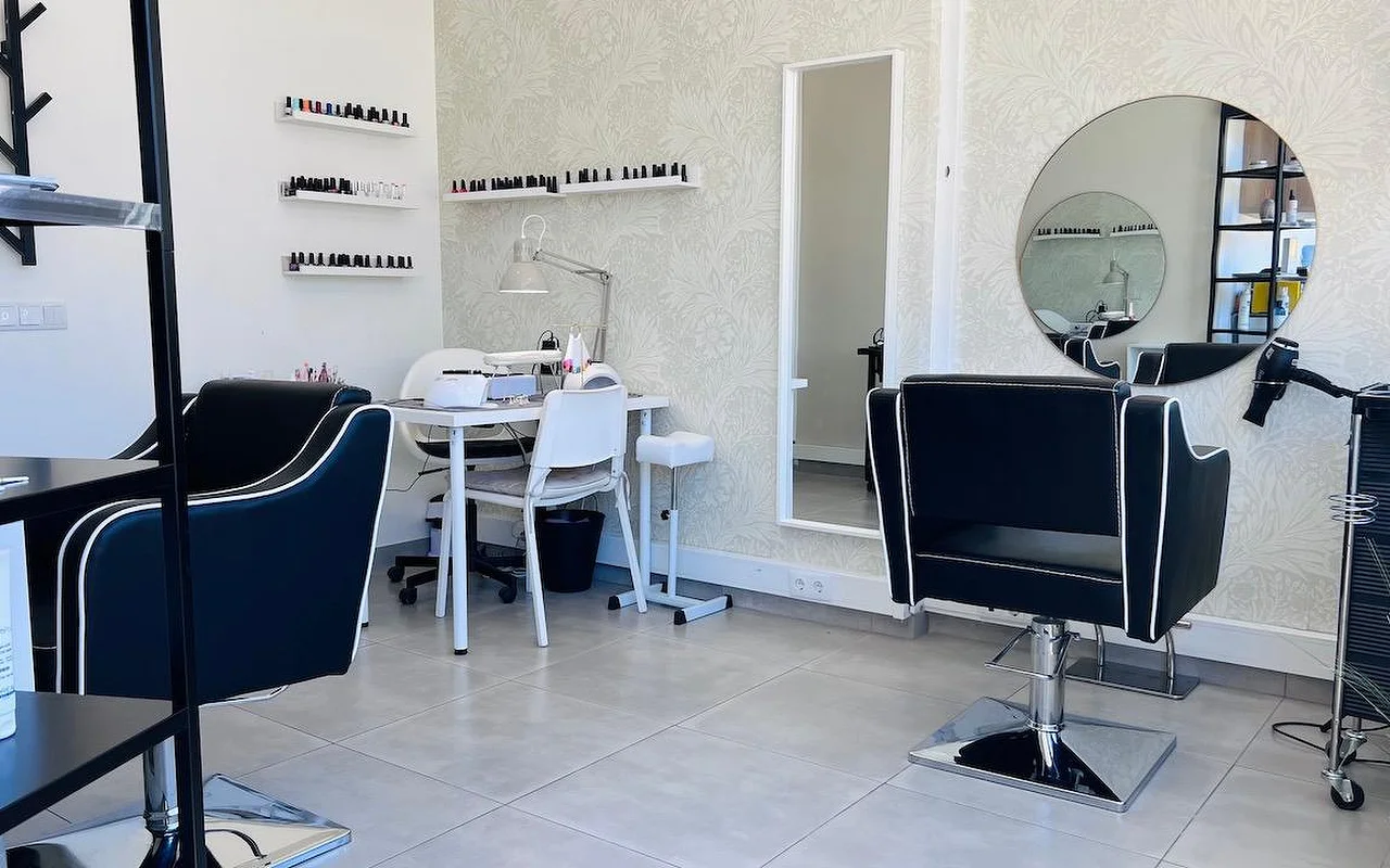 Ein kompakter Haar- und Nagelsalon mit schwarzen, weiss paspelierten Frisierstühlen, einem runden Spiegel und einem hohen Spiegel vor einer grauen Blümchentapete und Wandregalen voller Nagellacke.