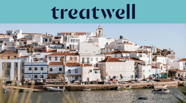 Ein weiss getünchtes Fischerdorf an der Algarve mit terrakottagedeckten Häusern, die einen Hügel über einem ruhigen Gewässer hinaufsteigen, kleinen Fischerbooten am Kai, Palmen und einem Kirchturm unter einem klaren blauen Himmel.