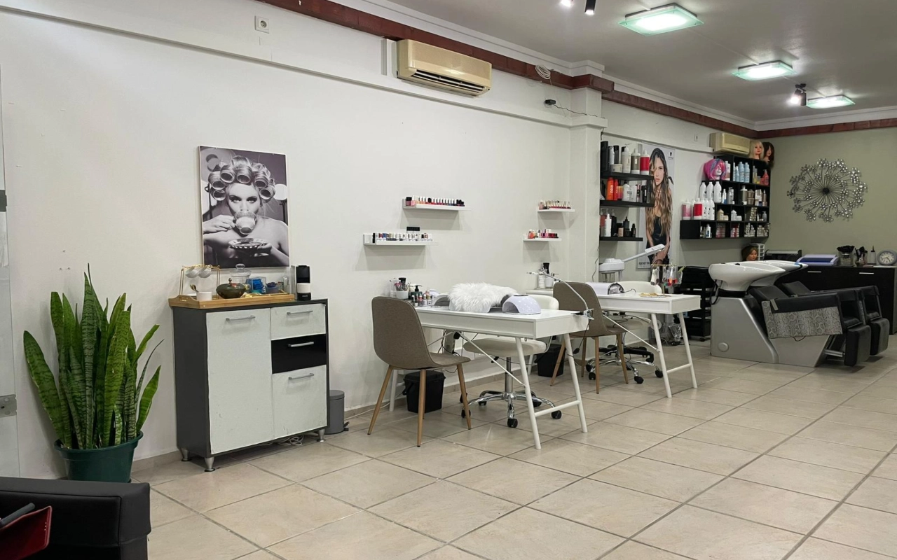 Ein Haar- und Beautysalon mit zwei weissen Maniküretischen und taupefarbenen Stühlen, einem Waschbecken und Produktregalen entlang der Wand, einem hohen Bogenhanf in der Ecke und einer grossen Schwarz-Weiss-Fotografie einer Frau mit Lockenwicklern.