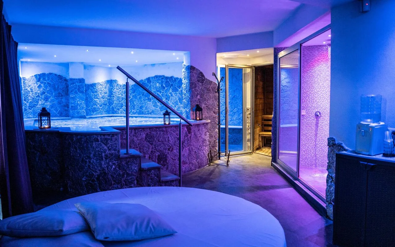 Eine private Spa-Suite bei Bellezza e Benessere in Taormina, in Blau und Violett beleuchtet, mit einem eingelassenen Whirlpool in einer Steinverkleidung, einer Holzsauna und dahinter einem mosaikgefliesten Dampfbad, und einem runden Bett im Vordergrund.