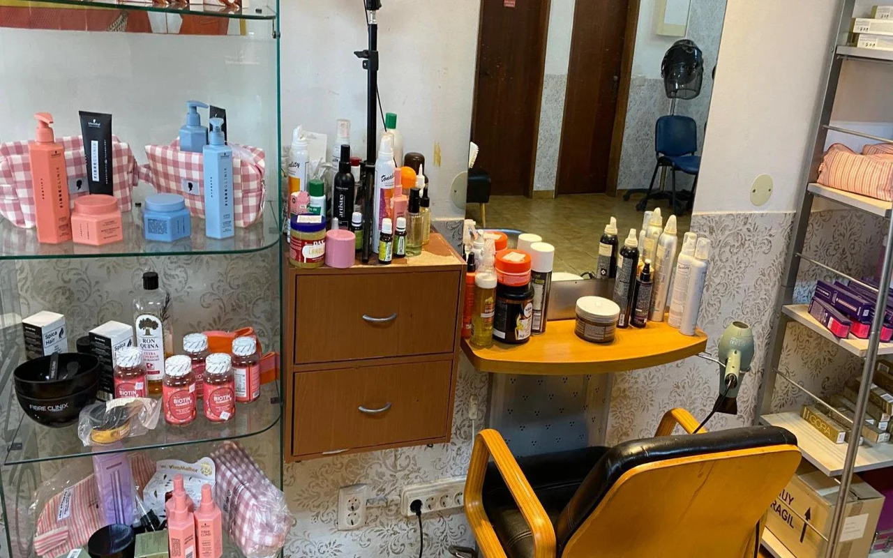 Der Arbeitsplatz einer Coiffeuse mit einem Wandspiegel und einer Holztheke voller Stylingprodukte, einer Glasvitrine mit Haarpflegeprodukten und Nahrungsergänzungsmitteln daneben und einem klassischen Frisierstuhl in Holz und Schwarz.