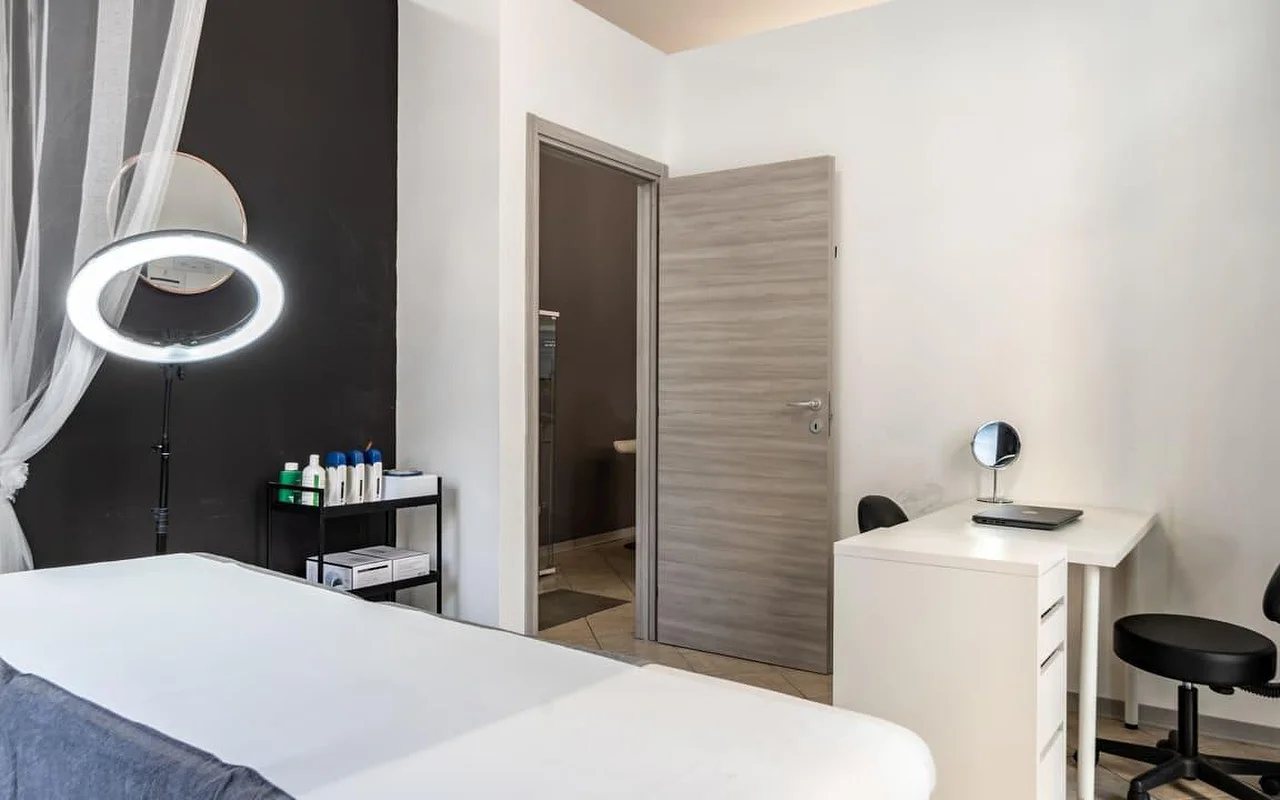 Ein Behandlungsraum bei Coquette Wellness and Beauty, mit einer weissen Liege, einem beleuchteten Ringlicht auf einem Ständer vor einer schwarzen Akzentwand, leichten Vorhängen am Fenster und einem kleinen weissen Schreibtisch mit Schminkspiegel neben einer Tür zu einem eigenen Bad.