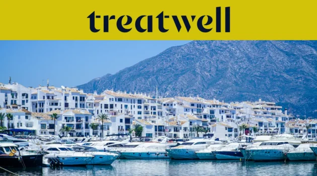 Der Yachthafen Puerto Banús in Marbella, gesäumt von weissen Apartmenthäusern und vertäuten Yachten vor Bergkulisse, unter einem gelben Treatwell-Banner.