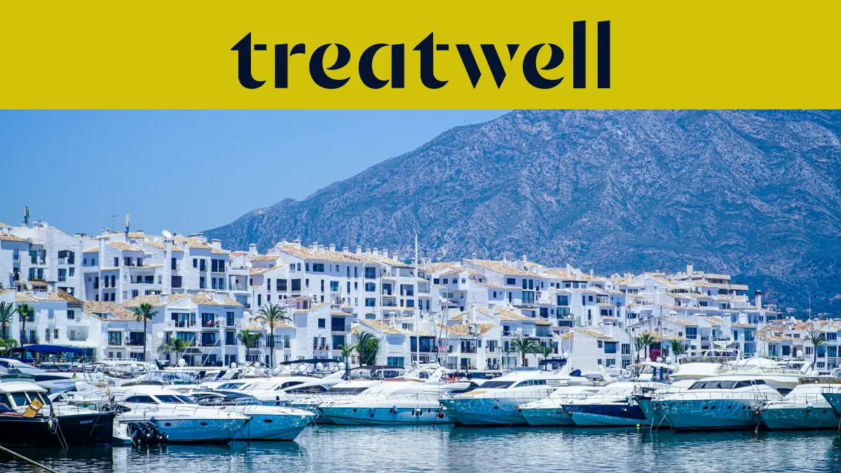 Der Yachthafen Puerto Banús in Marbella, gesäumt von weissen Apartmenthäusern und vertäuten Yachten vor Bergkulisse, unter einem gelben Treatwell-Banner.