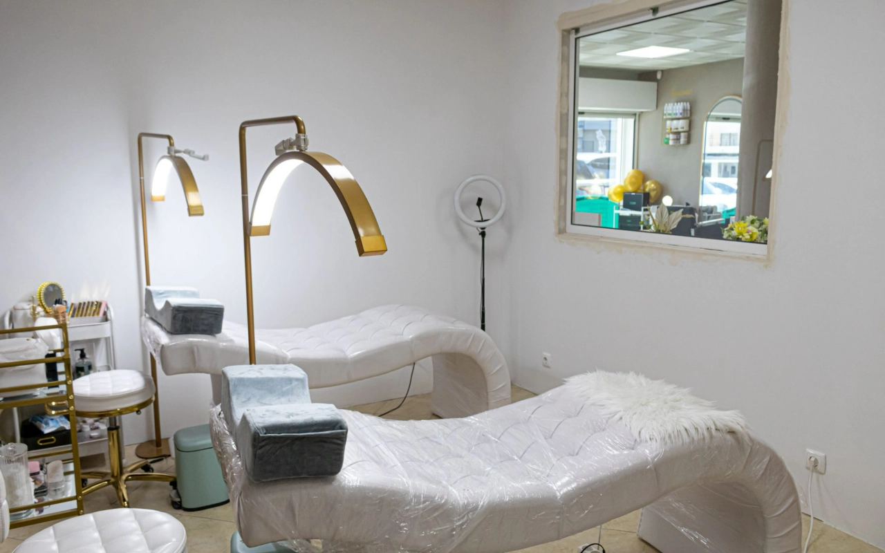 Ein heller Beauty-Behandlungsraum mit zwei weissen gesteppten Liegen, hohen goldenen Bogenlampen und einem Ringlicht, mit einem Innenfenster mit Blick zur Rezeption.