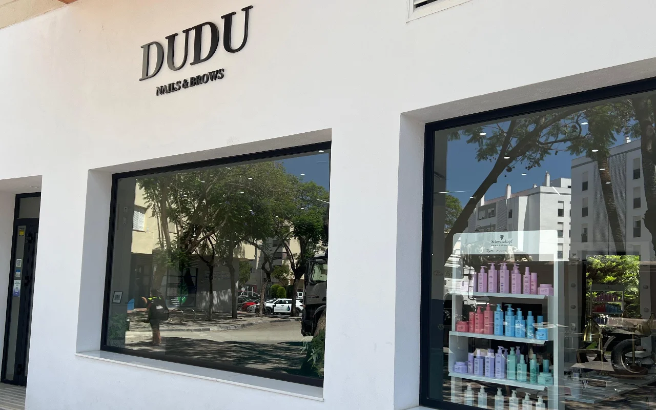Die weisse Ladenfront von Dudu Nails and Brows in Estepona, der Name in dunklen Lettern über breiten Schaufenstern mit Reihen von Haarpflegeprodukten.