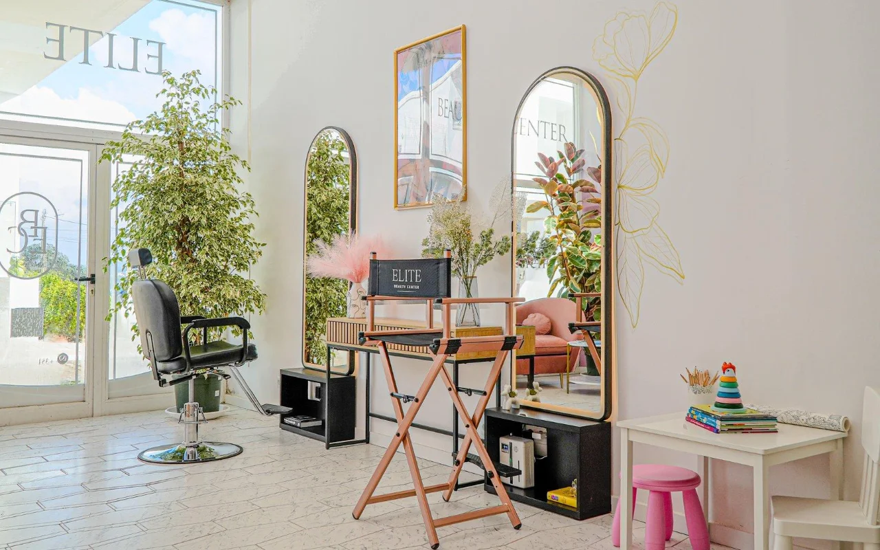 Ein heller, luftiger, ganz in Weiss gehaltener Beautysalon mit gewölbten goldgerahmten Spiegeln, einem schwarzen Frisierstuhl und einem klappbaren Regiestuhl, hohen grünen Pflanzen, rosa Akzenten und einer kleinen Kinderecke am Fenster.