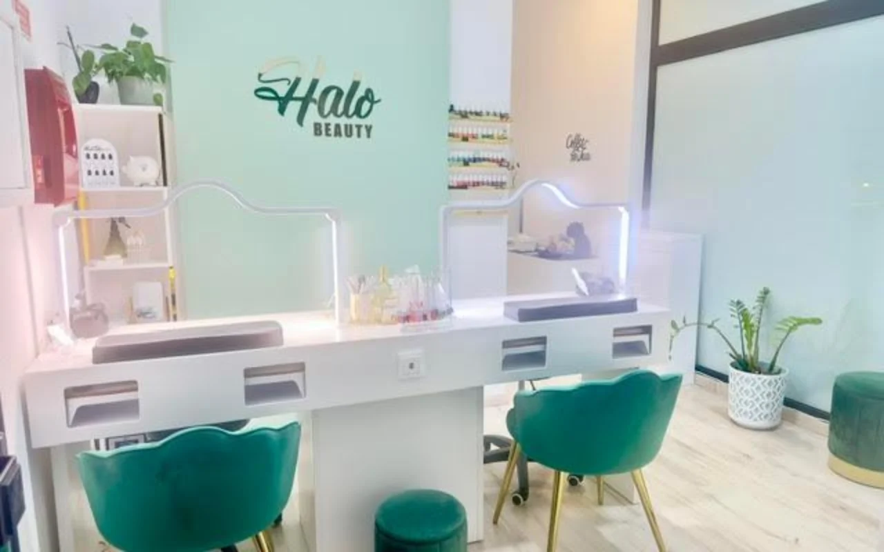 Ein Nagelstudio mit einer mintgrünen Akzentwand mit dem Halo-Beauty-Logo, einem weissen Doppel-Maniküretisch mit beleuchteten Plexiglas-Trennwänden, zwei smaragdgrünen Samt-Tonnensesseln, einem Wandregal voller Nagellacke und Topfpflanzen.