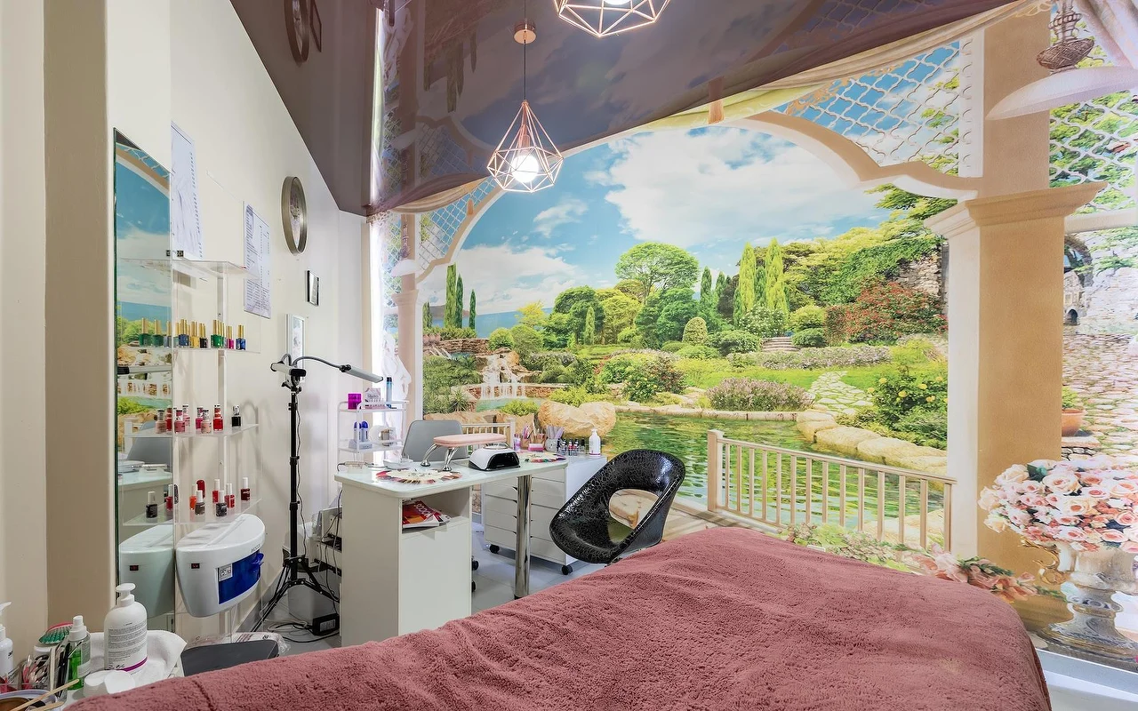 Ein Beauty-Behandlungsraum mit einer rosa drapierten Liege und einer weissen Nagelstation, vor einem grossen Wandbild mit Garten und See, mit Glasregalen voller Produkte daneben.