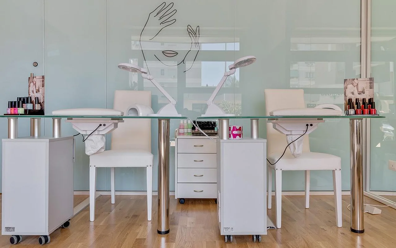 Ein Nagelstudio bei Perfetta Urban Beauty, mit zwei Maniküretischen mit Glasplatte und weissen Stühlen, Arbeitsleuchten, bunten Lack-Displays und einer mintgrünen Glaswand mit der Strichzeichnung eines Gesichts und zweier Hände.