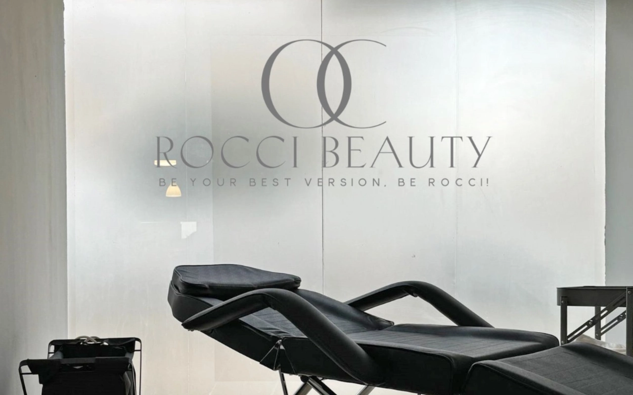 Ein Behandlungsraum mit einer schwarzen Liege zum Zurücklehnen und einem kleinen Gerätewagen, vor einer Milchglaswand mit dem Logo und dem Slogan von Rocci Beauty.