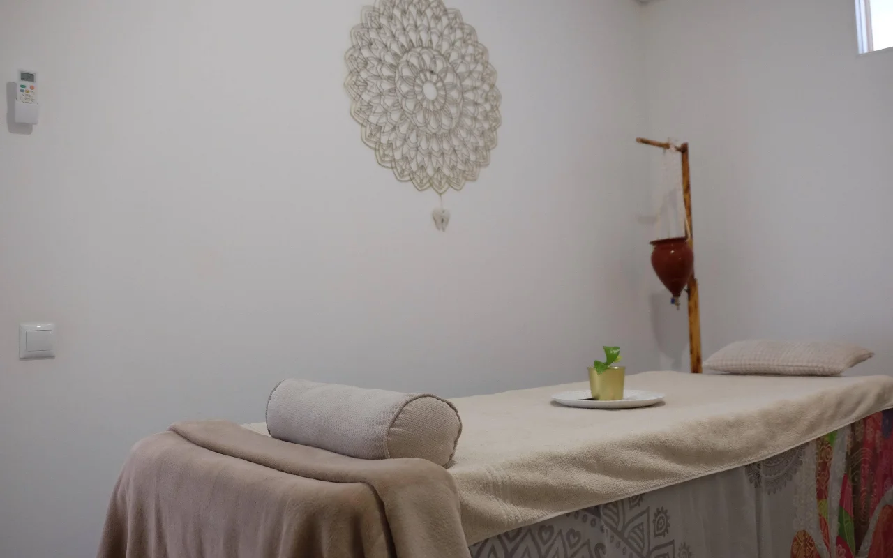 Ein ruhiger Massageraum in sanften, neutralen Tönen, mit einer mit einem Handtuch bedeckten Massageliege und einer Nackenrolle, einem grossen gehäkelten Mandala an der Wand und einem Holzständer, der einen hängenden Terrakottatopf hält.