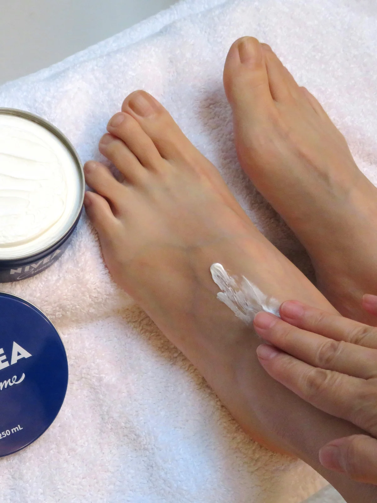 Nahaufnahme der Füsse einer Frau von oben, die auf einem weissen Tuch ruhen, daneben ein offener Tiegel Creme, während sie ihre Füsse eincremt